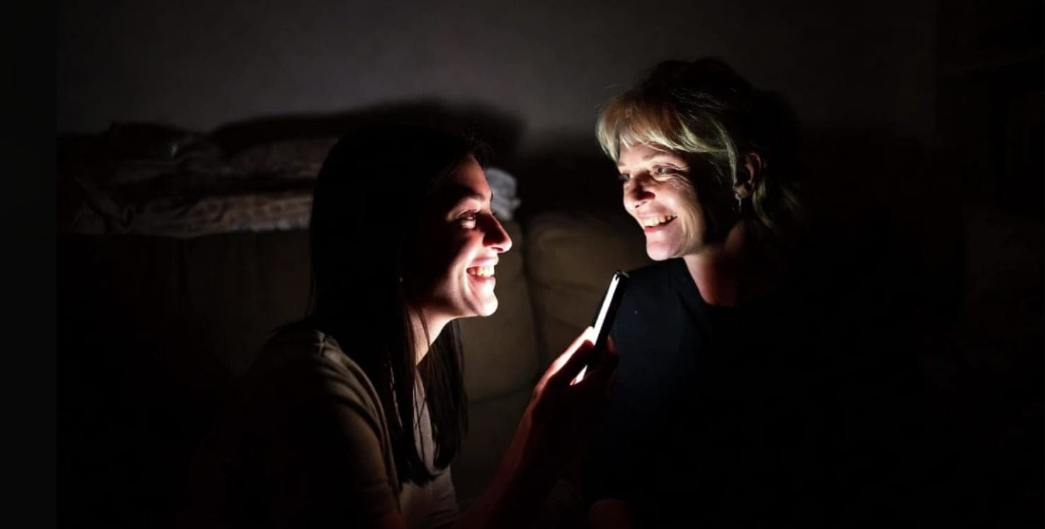 Two women sitting close in a dark room, both smiling at a phone held between them, their faces lit by the screen.